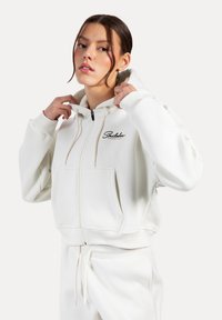 Witte ritsjack gemaakt van zacht materiaal, met een voorzak, trekkoordcapuchon en zwart logo op de linkerkant van de borst.