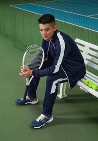 Marine sporttracksuit met witte strepen, blauwe tennisschoenen, houdend een tennisracket, zittend op een witte bank, tennisballe in de buurt.