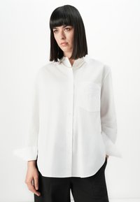 Woman with straight dark hair wearing a white button-up shirt with a chest pocket and black pants, standing against a plain white background.
