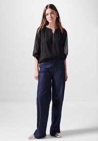 Schwarze, leichte Bluse mit einem Reißverschluss-Halsausschnitt; kombiniert mit lockeren, dunkelblauen Jeans; das Model trägt silberne flache Schuhe.