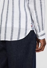 Camicia di lino bianca con righe verticali blu, con orlo curvo e polsini con bottoni; pantaloni in denim blu scuro visibili nella parte inferiore.