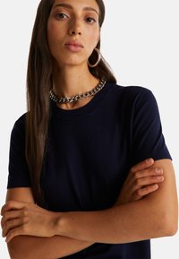 Námořnická bavlněná tričko s krátkým rukávem, kulatým výstřihem a jemnou texturou. Modelka má na sobě stříbrný řetízek a kruhové náušnice.