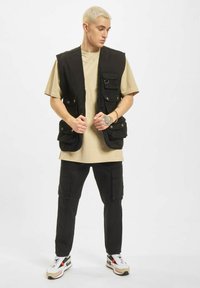 Svart ermeløs vest med flere lommer, brukt over en beige t-skjorte, kombinert med svarte cargobukser og flerfargede sneakers.