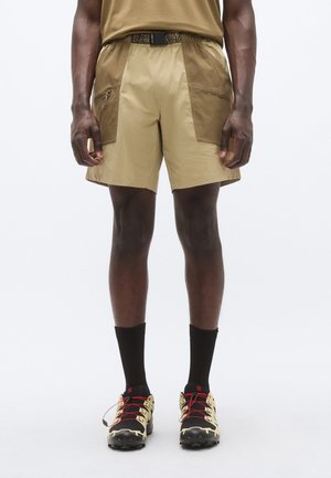 Beežid lühikesed püksid tumedamate toonide aktsentidega, tõmblukuga taskud, elastne vöökoht ning mustade sokkide ja must-kollaste vabaõhujalatsitega.