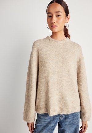 Pull en tricot beige avec un col rond, coupe décontractée, manches longues et ourlet côtelé. Porté sur un jean en denim bleu. Texture douce.