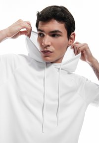 Biała krótkosz жұмыс in with a unique funnel neck, drawstrings, and textured fabric. Smooth surface with raglan sleeves.