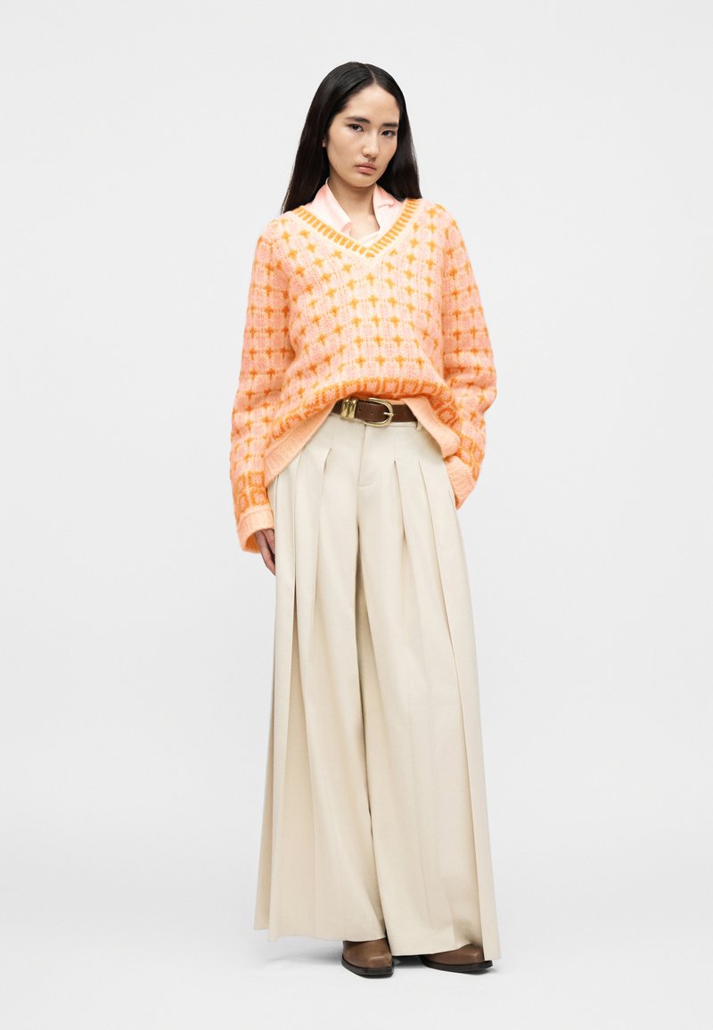 Femme portant un pull à motifs roses et oranges, un pantalon beige à jambes larges plissé, une ceinture marron et des chaussures marron, debout devant un fond blanc.