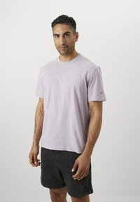 Helllila Baumwoll-T-Shirt mit kurzen Ärmeln, Rundhalsausschnitt und Logodetail auf der Brust. Kombiniert mit dunklen Shorts für einen lässigen Look.