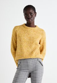 Un pull jaune texturé avec un col rond, associé à un pantalon à carreaux gris avec des poches zippées. Un matériau doux et duveteux est visible.