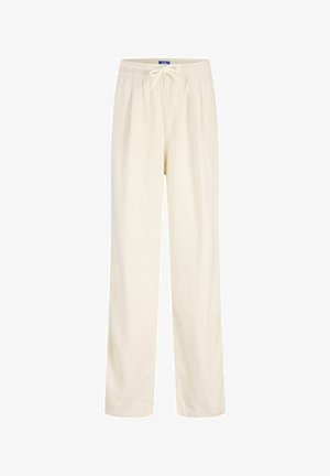 Ung person som står, iført en svart kortermet skjorte, løse hvite bukser og brune sko mot en ensfarget lys bakgrunn.