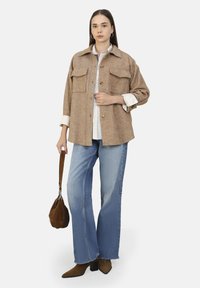 Bruine herringbone jas met twee borstzakken, eronder een wit shirt, gecombineerd met lichtblauwe flare jeans en bruine hoge laarzen.