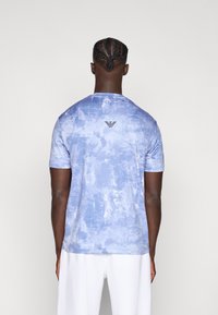 Ανοιχτό μπλε κοντομάνικο μπλουζάκι με υφασμένο μοτίβο tie-dye και μικρό μαύρο λογότυπο στο πάνω μέρος της πλάτης. Μαλακό ύφασμα, στρογγυλή λαιμόκοψη.