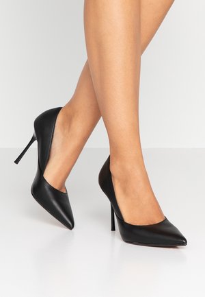 Piernas con tacones altos negros de punta afilada y tacón de aguja, sobre una superficie blanca lisa con un fondo gris claro.