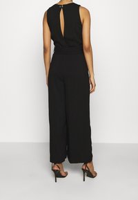 Černý bezramenný overal s detailem ve tvaru klíčové dírky na zádech, širokými nohavicemi a přiléhavým pasem. Vyroben z hladké látky s matným povrchem.