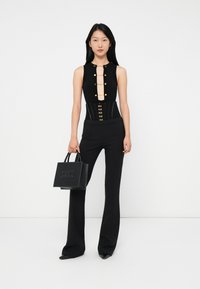Woman in black sleeveless top with gold buttons, high-waist pants, holding a black handbag, standing against a white background.