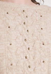Lichtbeige gebreide trui met kabelpatronen en subtiele gouden metalen accenten die gelijkmatig over de voorkant zijn verdeeld. Zachte textuur.
