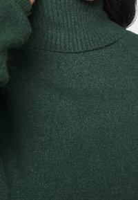 Zielony sweter z golfem, wykonany z dzianiny, o prążkowanym dekolcie i miękkiej fakturze, przedstawiający zbliżenie na wzór dzianiny.