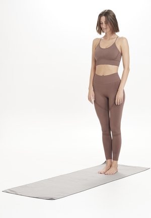 Brown athletic crop top and leggings on a person standing barefoot on a gray yoga mat. Smooth fabric and fitted design, seamless details.
