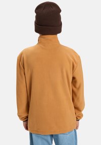 Pull en polaire couleur beige chaud avec col haut, manches longues et coupe décontractée. Accompagné d'un bonnet tricoté marron foncé.