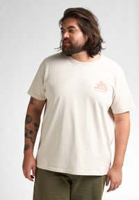 Beige katoenen t-shirt met een ronde hals, korte mouwen en een subtiele roze grafische logo op de linkerborst. Zachte textuur, relaxte pasvorm.