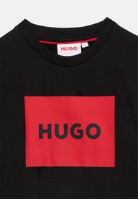 Μαύρο φούτερ με στρογγυλή λαιμόκοψη και κόκκινο ορθογώνιο λογότυπο που δείχνει το όνομα της μάρκας "HUGO" με έντονα μαύρα γράμματα.