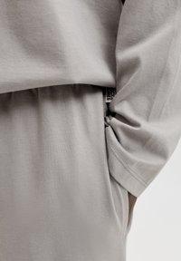 Chemise oversize gris clair avec des manches amples et un détail froncé sur le côté. Texture en coton doux avec un léger éclat.