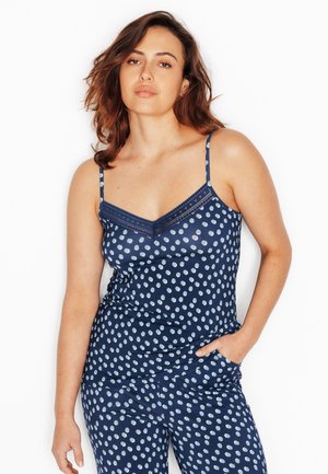 Vrouw met bruin schouderlang haar, gekleed in een marineblauw mouwloos topje met witte stippen en bijpassende broek, poseert met één hand in de zak.