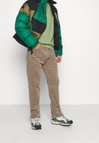 Doudoune en vert, noir et olive ; t-shirt vert clair ; pantalon en velours côtelé beige ; baskets noires et vertes avec des accents jaunes.
