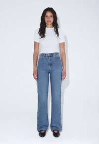 Jeans de mezclilla azul de tiro alto y acampanados con costuras visibles. Combinados con una camiseta blanca lisa y zapatos negros. La modelo está de pie contra un fondo blanco.