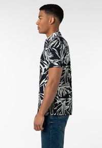 Schwarz kurzärmliges Hemd mit weißem botanischen Druck, mit Knöpfen und klassischem Kragen. Getragen mit blauen Jeans.