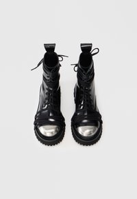 Čierne členkové topánky s lesklým koženým zvrškom, gumovou podrážkou a strieborným špičkovým krytom. Obsahujú zložitú šnurovanie a textúrované popruhy, ktoré obopínajú topánku.