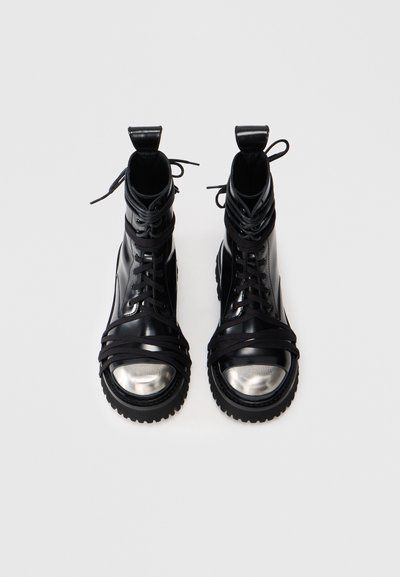 Stivaletti neri alla caviglia con tomaia in pelle lucida, suola in gomma e punta in metallo argentato. Presentano un complesso sistema di allacciatura e fascette testurizzate che si avvolgono attorno.