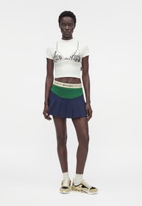 Γυναίκα που φοράει λευκό crop top με στάμπα σουτιέν, πράσινη και μπλε πλισέ μίνι φούστα, λευκές κάλτσες και μπεζ-μαύρα αθλητικά παπούτσια, στέκεται μπροστά σε λευκό φόντο.