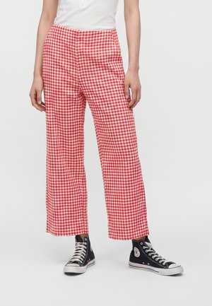Person iført rød- og hvidternede bukser med brede ben, sorte høje sneakers og en hvid skjorte, står mod en ensfarvet baggrund.