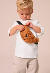 Brun plysch rund väska med ett björnfacedesign, svart dragkedja och justerbar svart rem; barn som bär en vit långärmad skjorta och beige byxor.