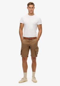 Bruine cargoshorts met meerdere zakken, gedragen met een aansluitend wit T-shirt en een bruine riem. Beige sneakers en lichtgekleurde sokken maken de outfit af.