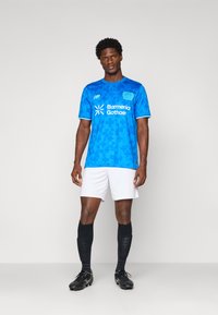 Male athlete standing wearing a blue patterned soccer jersey, white shorts, black knee-high socks, and black cleats against a plain background.