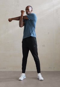 Idrettsantrekk med en tettsittende blå kortermet skjorte, svarte tilpassede bukser og hvite joggesko med blå detaljer. Glatt, pustende stoff.