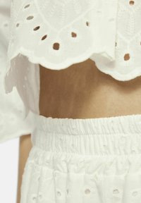 Witte top met golfzoom en geborduurde uitsparingen, gecombineerd met witte, gathered elastische tailleband onderkant met textuurdetails.