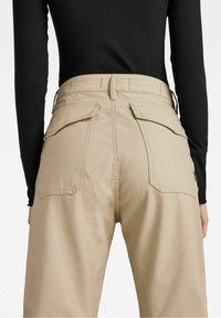 Krémové kalhoty s vysokým pasem a velkými kapsami na flap vzadu, nošené s černým dlouhým rukávem s žebrováním, zobrazené zezadu.