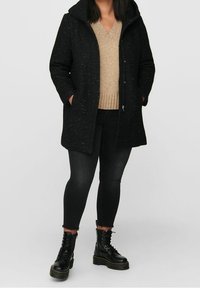 Abrigo negro texturizado con capucha, cremallera frontal y bolsillos. Llevado sobre un suéter beige y combinado con jeans ajustados negros y botas con cordones.