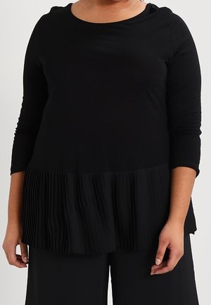 Vrouw draagt een zwarte top met lange mouwen met een geplooide peplumzoom en bijpassende zwarte broek, staand tegen een effen achtergrond.