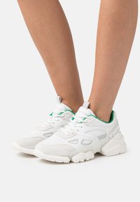 Scarpe sportive bianche con pannelli in rete e dettagli verdi. Presentano una suola spessa e un sistema di allacciatura prominente, combinando texture e design moderno.