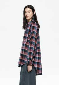 Camisa de franela a cuadros en rojo y negro con mangas largas, dobladillo curvado, un bolsillo en el pecho y diseño en capas. Se lleva con una falda gris.