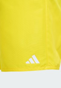 Žlté športové šortky s textúrovaným povrchom, s bielym logom Adidas blízko lemu a jemnými detailmi šitia.