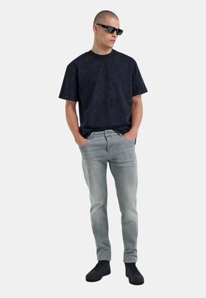 Camiseta de manga corta oversized en azul marino oscuro, jeans ajustados en gris claro y zapatillas negras. El modelo lleva gafas de sol negras y está de pie con las manos en los bolsillos.