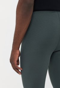 Tummanvihreät ribbileggingsit, joissa on tiukka muotoilu, esiteltynä käden levätessä kankaan päällä. Tekstuurin näyttäytyy sileänä ja joustavana.
