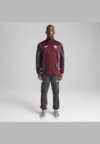 Bordeaux træningsdragt med grå accenter, bestående af en jakke med lynlås og bukser. Jakken har en høj krave og logoer på begge dele. Grå sneakers.
