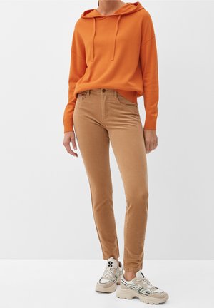 Persoon die een oranje capuchontrui, beige slanke broek en witte chunky sneakers draagt, tegen een effen lichtgrijze achtergrond.