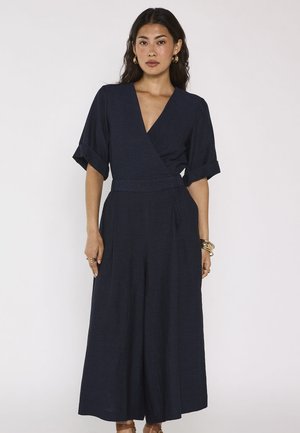 Femme portant une combinaison bleu marine style portefeuille avec jambes larges et manches mi-longues, accessoirisée avec des boucles d'oreilles créoles dorées et des bracelets.